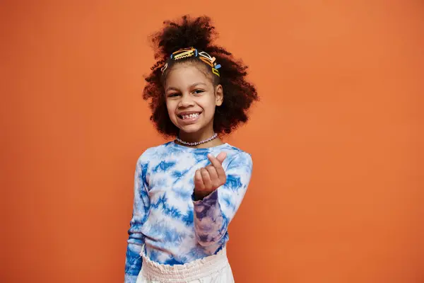A young African American girl with curly hair and hair clips smiles at the camera while wearing a blue tie-dye shirt. — Stock Photo