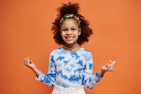 Young African American girl smiling in blue and white tie-dye shirt and hair clips on orange background. — Stock Photo
