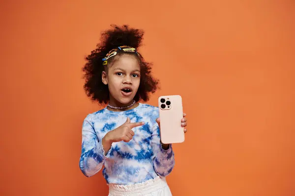 Young African American girl with colorful hair clips stands in trendy outfit against orange background, holding a phone and pointing to it. — Stock Photo