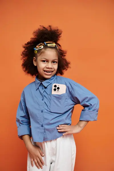 A young girl with a trendy hairstyle stands on an orange background, looking confident and stylish in a blue shirt with smartphone in a pocket — Stock Photo
