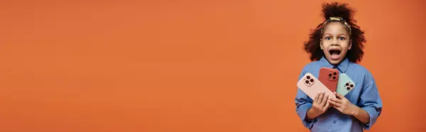 A young African American girl with a trendy hairstyle holds up three phones in front of an orange background. — Stock Photo