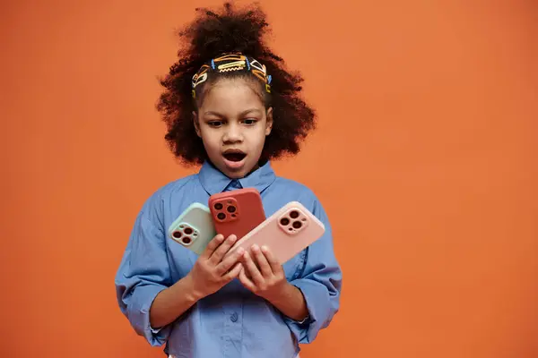 A stylish African American girl holds three phones against an orange background with a surprised look. — Stock Photo