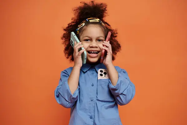 Young black girl with stylish hair clips, smiling playfully against an orange backdrop, holding two phones to her ears. — Stock Photo