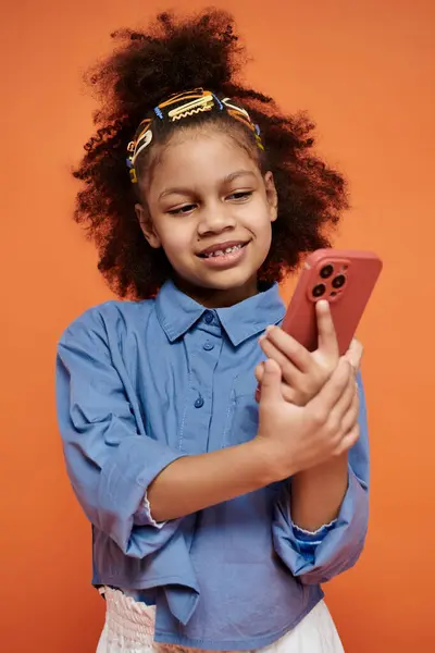 A young girl with a stylish outfit and hair clips smiles while holding a smartphone against an orange background. — Stock Photo