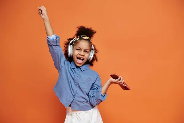 Une jeune fille élégante avec des pinces à cheveux danse avec excitation tout en portant un casque et en tenant un téléphone sur un fond orange. — Photo de stock