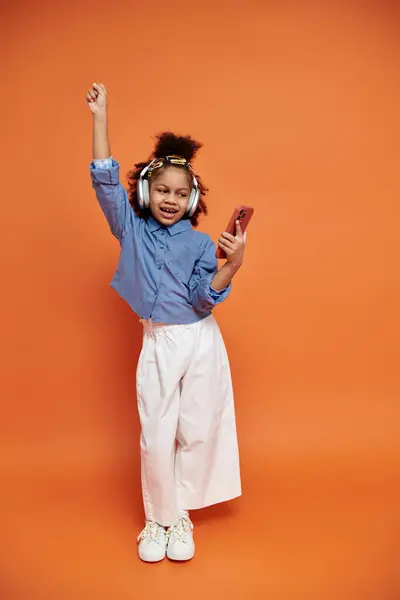 Чорна дівчина в модному одязі радісно танцює з навушниками та телефоном на помаранчевому фоні. — стокове фото