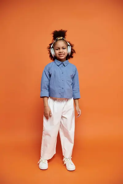Confident young African American girl in chic outfit stands against orange background, styled hair, radiating confidence. — Stock Photo