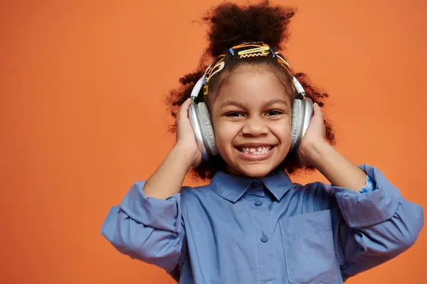 A young African American girl with a big smile, wearing a blue shirt, headphones, and hair clips, poses in front of an orange background. — Stock Photo