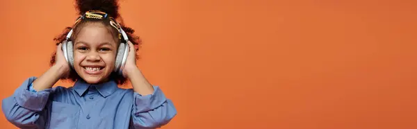 A young girl with a stylish outfit and hair clips smiles while wearing headphones against a bright orange backdrop. — Stock Photo