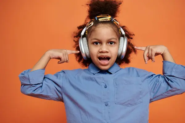 Une afro-américaine avec des pinces à cheveux porte un casque et une tenue élégante tout en posant sur un fond orange vif. — Photo de stock