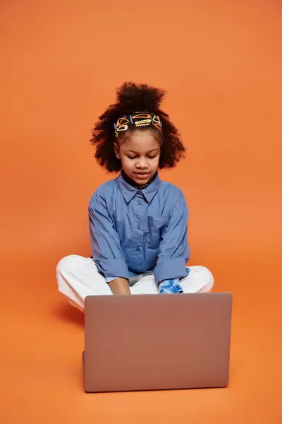 An African American girl with stylish hair clips sits on an orange background, focused on her laptop. — Stock Photo
