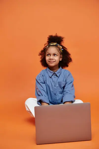 Une jeune afro-américaine avec des pinces à cheveux à la mode s'assoit sur un fond orange, à l'aide d'un ordinateur portable. — Photo de stock