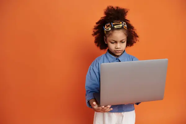 Stylish African American girl in trendy outfit stands in front of orange background, hair in ponytail with clips, holding a laptop. — Stock Photo