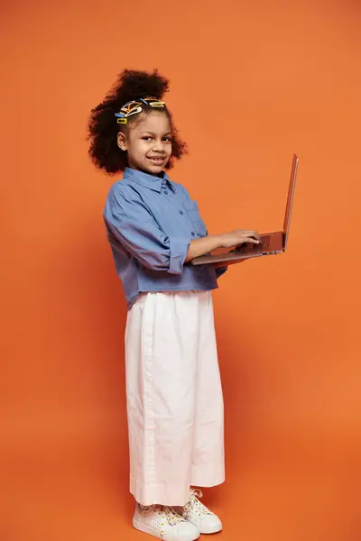 Ein junges Mädchen mit bunten Haarspangen steht in einem schicken Outfit und lächelt, während sie einen Laptop vor orangefarbenem Hintergrund benutzt.. — Stockfoto