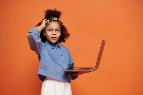 A stylish African American girl with hair clips, trendy outfit, holds a laptop against an orange backdrop. — Stock Photo