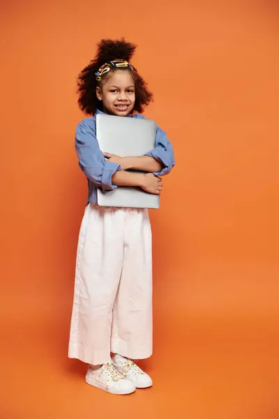 A young African American girl in a trendy outfit and hair clips stands confidently against an orange backdrop, holding a laptop. — Stock Photo
