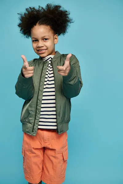 An adorable Black girl in a green jacket, striped shirt, orange shorts, smiles and points with both hands on a light blue backdrop. — Stock Photo