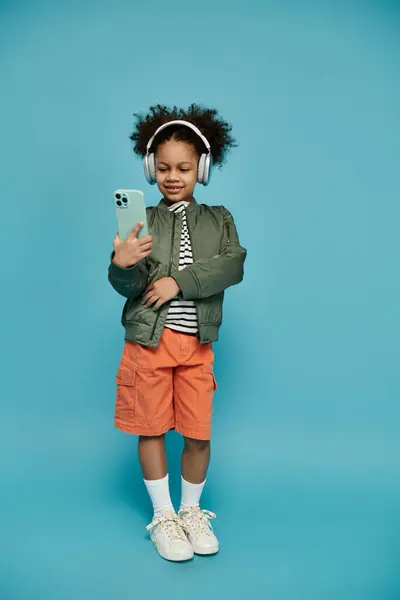 A young African American girl with curly hair is engrossed in her smartphone, headphones on, against a bright blue backdrop. — Stock Photo