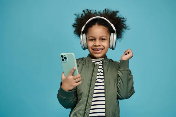 A young African American girl with curly hair smiles while wearing headphones and holding a smartphone against a blue background. — Stock Photo