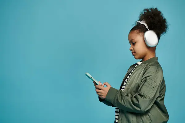 A young African American girl with curly hair wears headphones and uses a phone against a blue background. — Stock Photo