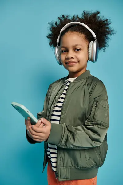 A young African American girl with curly hair smiles while wearing headphones and holding a phone in front of a blue background. — Stock Photo