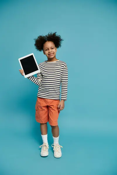 A young African American girl with curly hair smiles while holding a tablet in front of a blue background. — Stock Photo