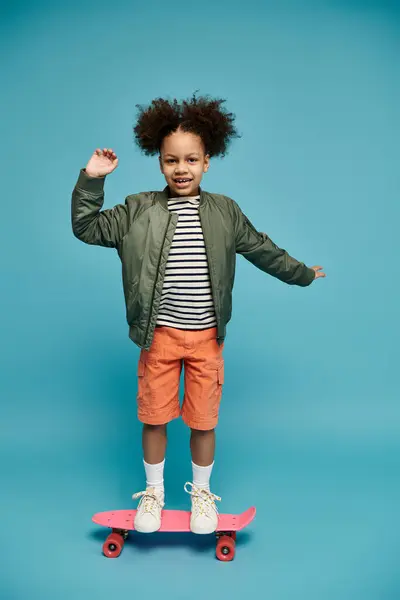 A young African American girl with curly hair smiles while standing on a pink skateboard against a blue background. — Stock Photo