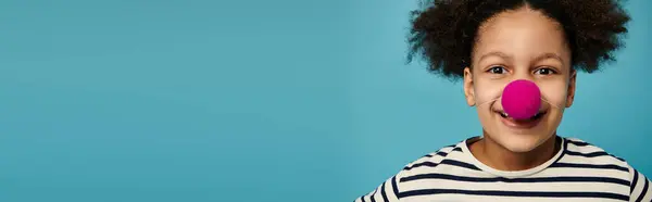 A young African American girl with curly hair wears a pink clown nose and smiles at the camera. — Stock Photo