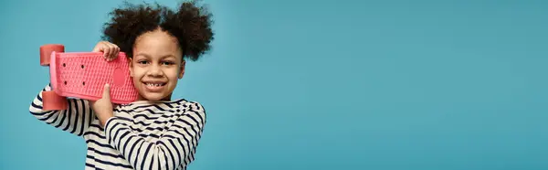 Une jeune afro-américaine aux cheveux bouclés sourit à la caméra tout en tenant une planche à roulettes rose sur un fond bleu. — Photo de stock