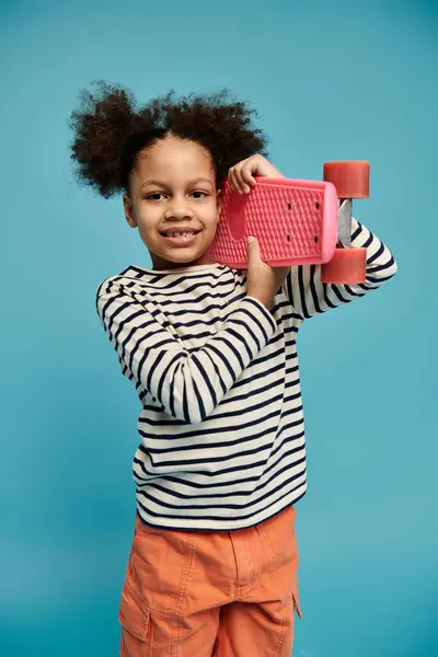 A young African American girl with curly hair smiles brightly as she holds a pink skateboard against a blue background. — Stock Photo