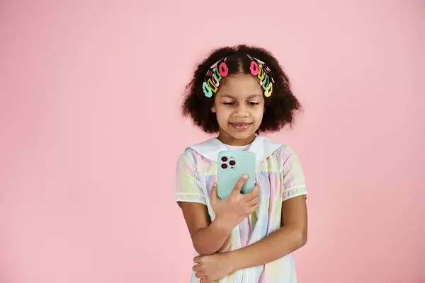A young African American girl with colorful hair clips stands on a pink background, smiling as she looks at a smartphone. — Stock Photo