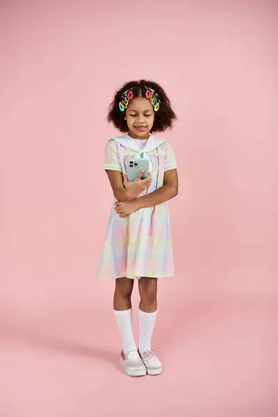 A young African American girl with colorful hair clips stands in a cute dress on a pink background. — Stock Photo
