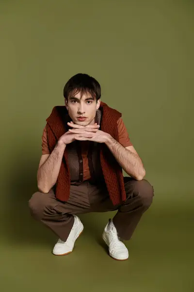 A man in a brown sweater and pants sits with his hands resting on his chin against a green backdrop. — Stock Photo