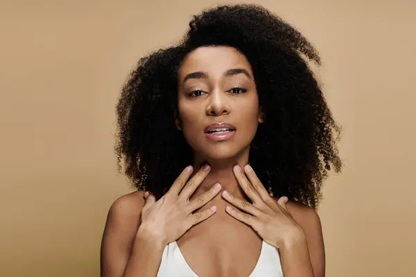 A beautiful African American woman with natural makeup poses against a beige background, her hands gently touching her neck. — Stock Photo