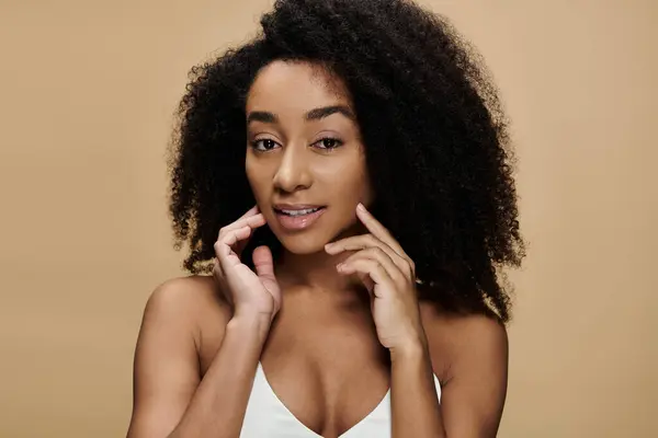 Uma bela mulher afro-americana com maquiagem natural posa contra um fundo bege, com as mãos suavemente tocando seu rosto. — Fotografia de Stock