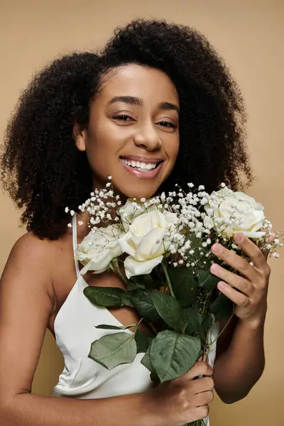 An African American woman with natural makeup smiles brightly while holding a bouquet of white roses against a beige background. — Stock Photo