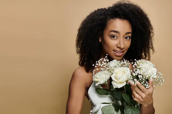 A beautiful African American woman with natural makeup holds a bouquet of white roses against a beige backdrop. — Stock Photo
