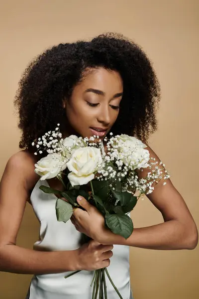 Une femme afro-américaine au maquillage naturel tient un bouquet de roses blanches et de souffle de bébé sur un fond beige. — Photo de stock