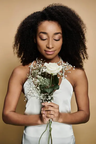 Una hermosa mujer afroamericana en un vestido de deslizamiento blanco sostiene un ramo de flores blancas mientras las mira. - foto de stock