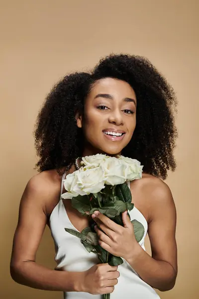 A beautiful African American woman with natural makeup smiles brightly while holding a bouquet of white roses against a beige background. — Stock Photo