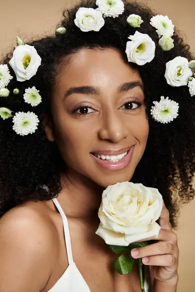 An African American woman with natural makeup smiles brightly, adorned with flowers in her hair and holding a white rose. — Stock Photo