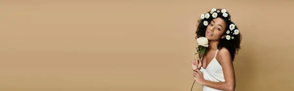 A beautiful African American woman with natural makeup and flowers in her hair poses against a beige backdrop, holding a white rose. — Stock Photo