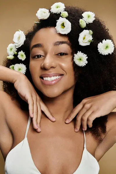 Una hermosa mujer afroamericana con maquillaje natural y flores blancas en su cabello sonríe brillantemente sobre un fondo beige. - foto de stock