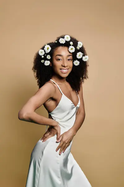 Uma bela mulher afro-americana em um vestido branco com flores em seu cabelo, sorri para a câmera contra um pano de fundo bege. — Fotografia de Stock