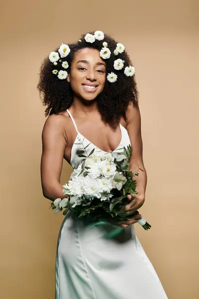Donna afroamericana sorridente in abito bianco, capelli fioriti, mazzo di fiori bianchi, posa davanti a sfondo beige. — Foto stock