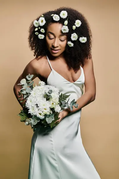 A beautiful African American woman with natural makeup and flowers in her hair holds a bouquet in front of a beige backdrop. — Stock Photo