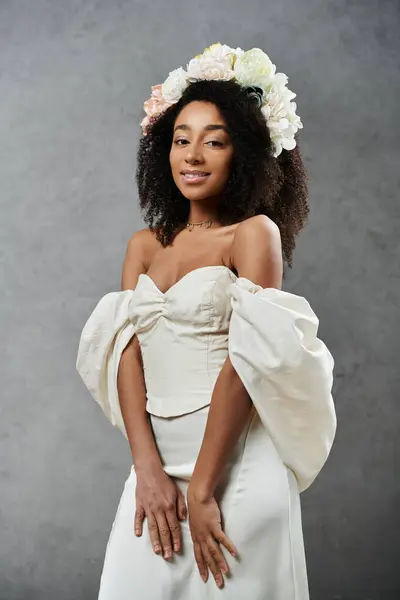 Eine schöne afroamerikanische Braut in einem weißen Hochzeitskleid und einer floralen Krone lächelt vor grauem Hintergrund. — Stockfoto