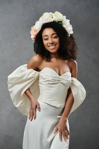 A beautiful African American bride in a white wedding dress with flowers in her hair smiles against a grey backdrop. — Stock Photo