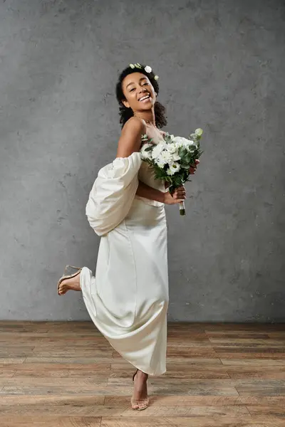 Una bella sposa afroamericana in un abito da sposa bianco e fiori tra i capelli sorride radiosamente su uno sfondo grigio. — Foto stock