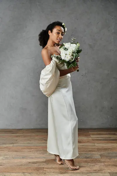 A beautiful African American bride stands in a white wedding dress and holds a bouquet of white flowers. — Stock Photo